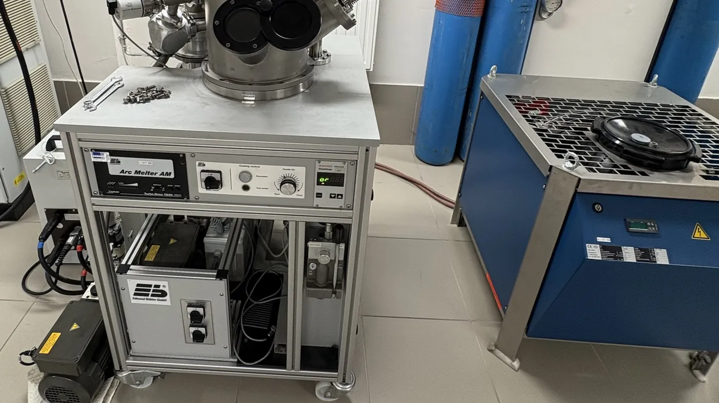 An arc melter in a university materials lab: the steel melting chamber with two round windows on its control cabinet, a chiller and gas bottles beside it.