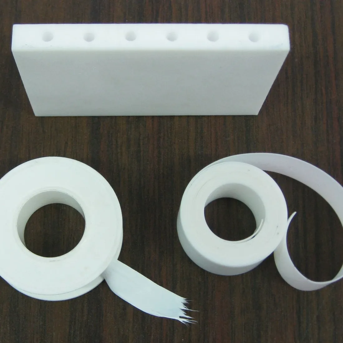 Ein weißer PTFE-Block mit gebohrten Löchern und zwei Rollen PTFE-Band auf einem Holztisch.