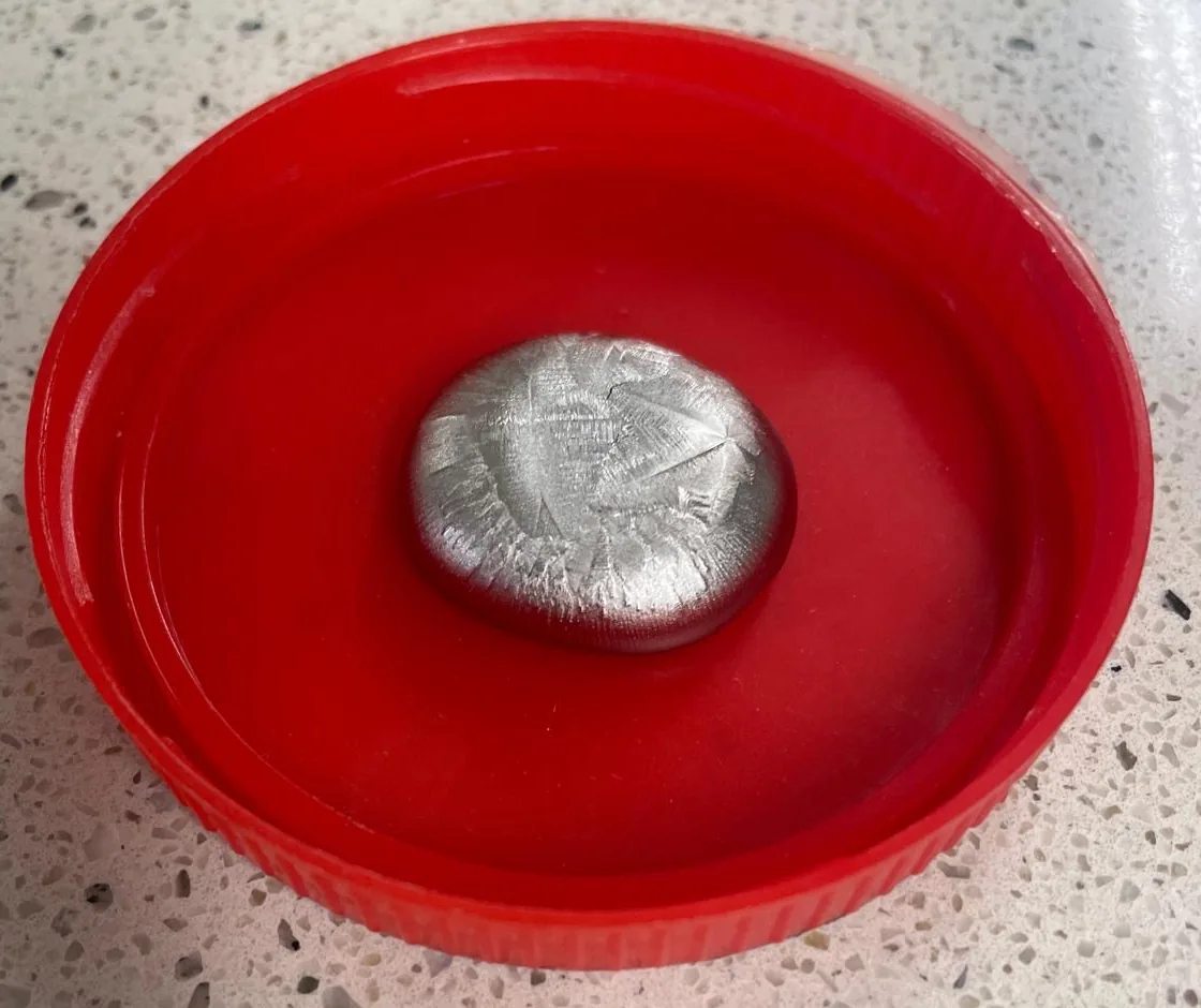 A cast alloy button with a crystalline surface, resting in a red lid on a lab bench.