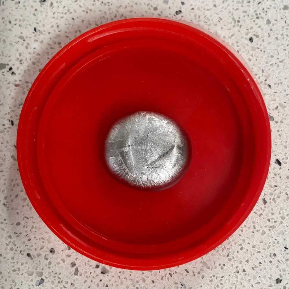 Ein gegossener Legierungsknopf mit kristallinem Oberflächenmuster, in einem roten Deckel auf einem Labortisch.